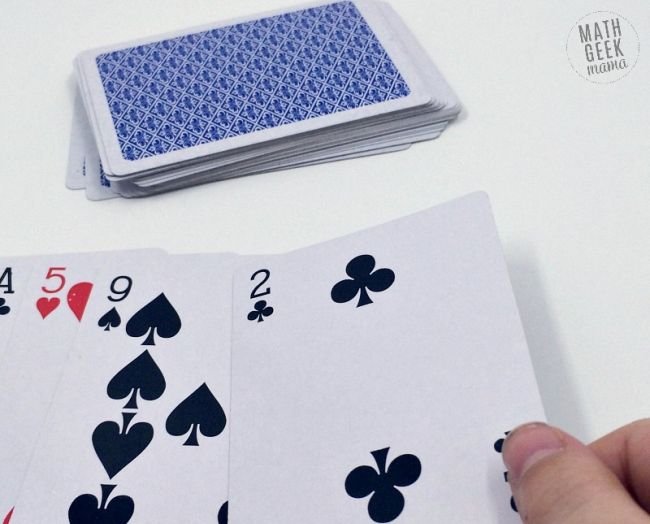 A hand holding a two of clubs as part of a set of cards turned face up next to a stack of cards piled up face down as an example of pi day activities