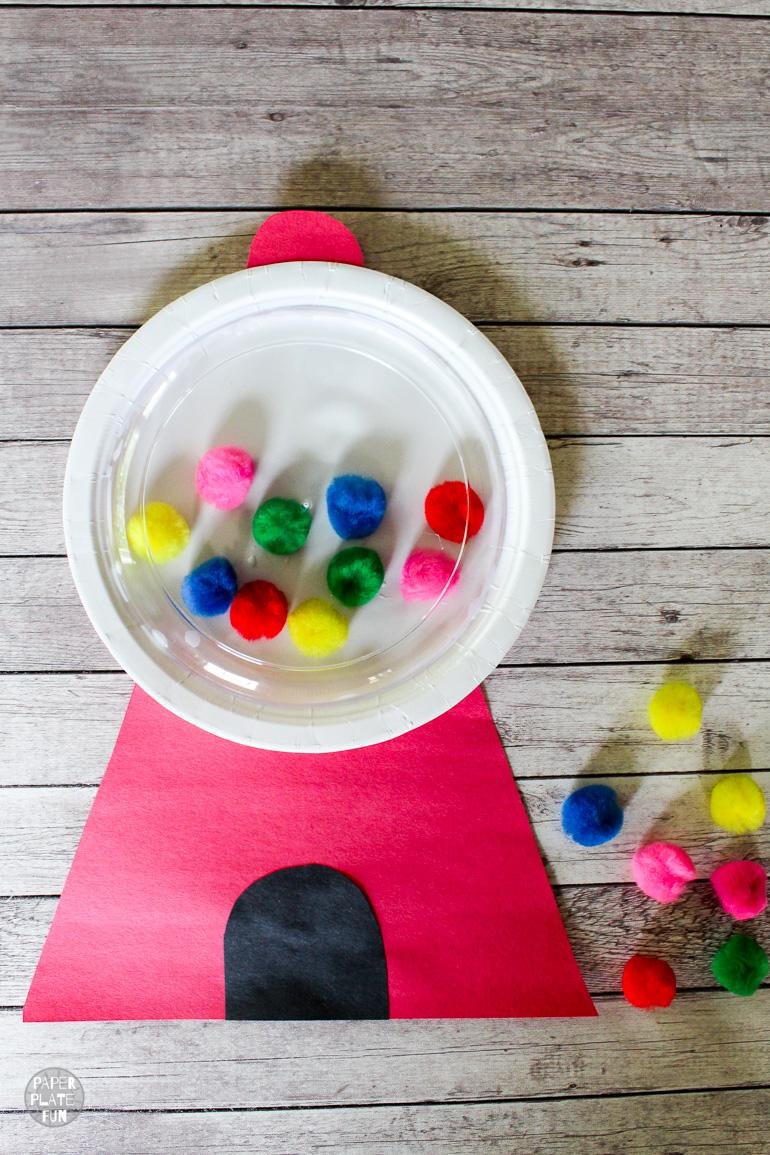 A white paper plate has different colored pom poms glued to it. Red cardstock completes the rest of the gumball machine 