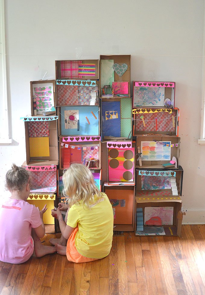 Two children sit in front of a collaborative art project that consists of decorated cardboard boxes attached to a wall.