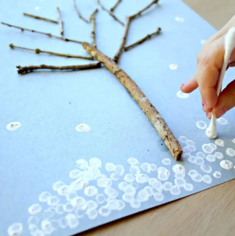Twigs glued to paper to form a bare winter tree, while a kindergartener paints snowflakes with a cotton swab
