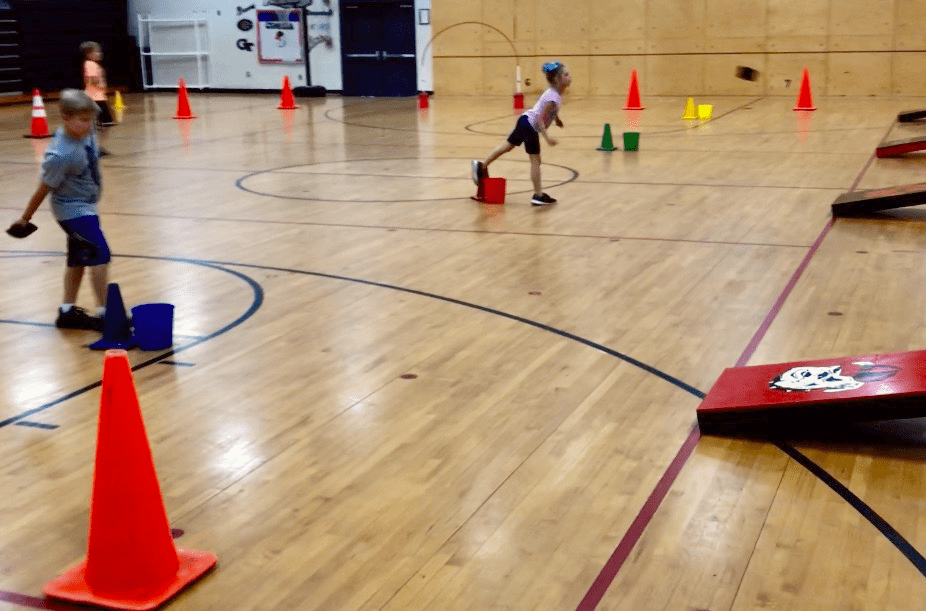Students stand about 10 yards back from cornhold boards. There are cones scattered throughout the gymnasium as an example of elementary PE games