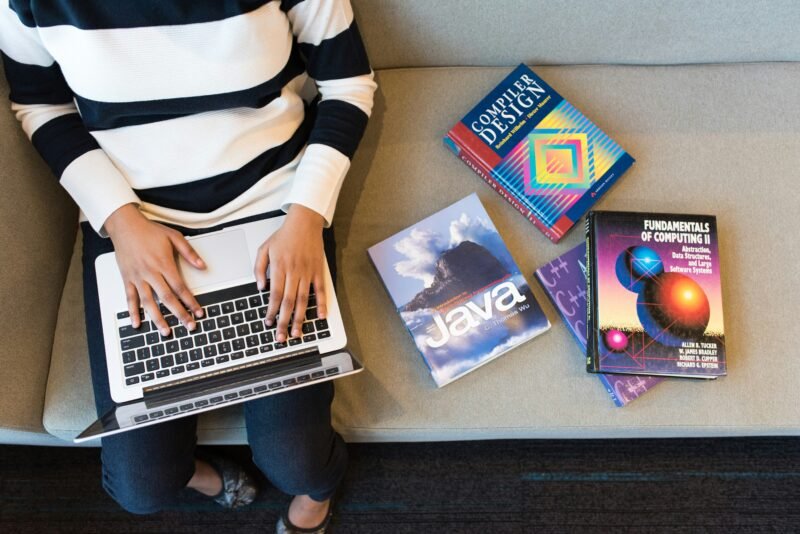A database administrator sits on a bench working on a laptop next to a pile of computer programming books