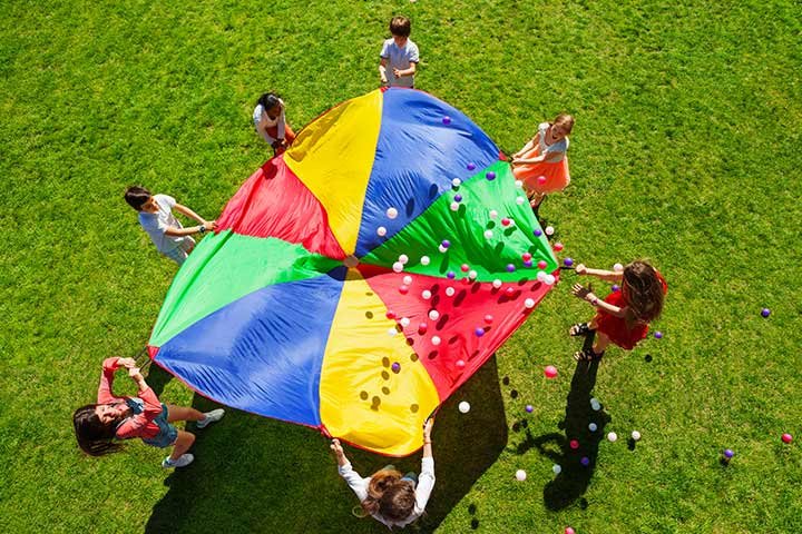 Students stand around a large parachute with small balls bouncing on the top of it as an example of elementary PE games