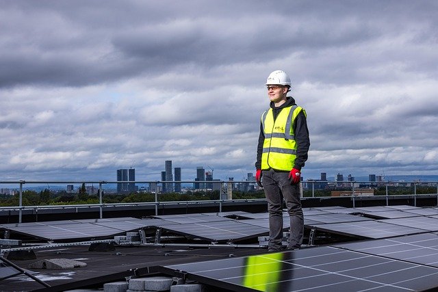 A Solar Photovoltaic Technician stands on a rooftop covered in solar panels, silhouetted against a city skyline