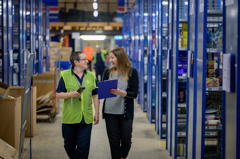 A store manager and employee look at a clipboard as they walk down the aisle of a warehouse