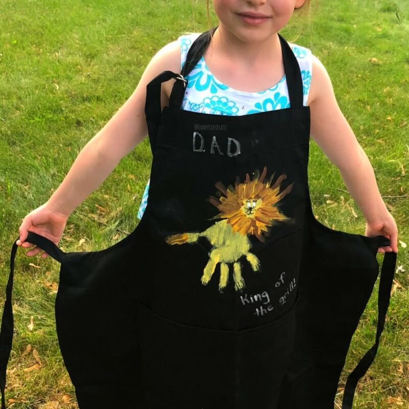 A child is seen wearing a black apron with a handprint design on it 