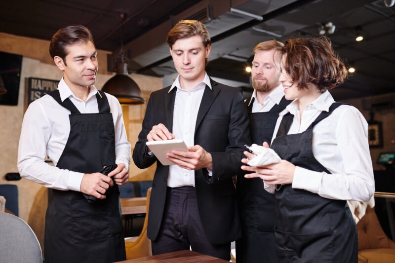 Serious confident handsome young restaurant manager using tablet while dividing responsibilities among waiters in modern establishment