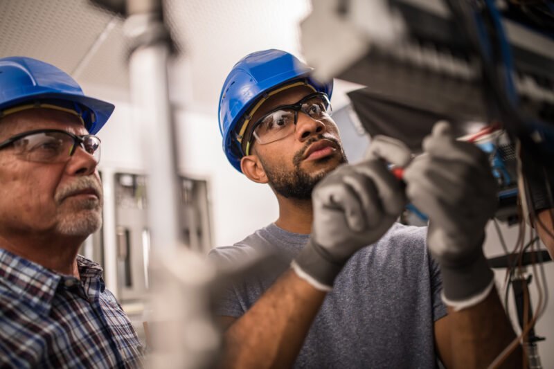 Photo of a senior caucasian and young electrician working on a fuse box.