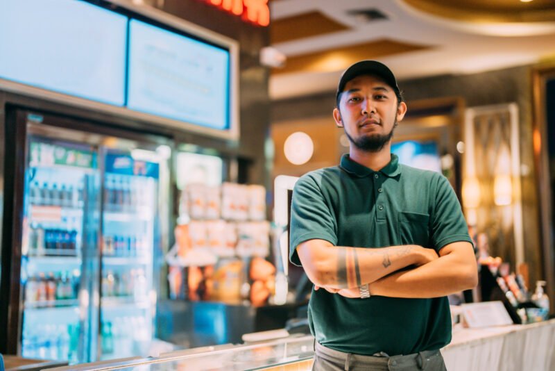 Portrait of tattooed cinema staff man against popcorn counter. He's wearing a uniform.