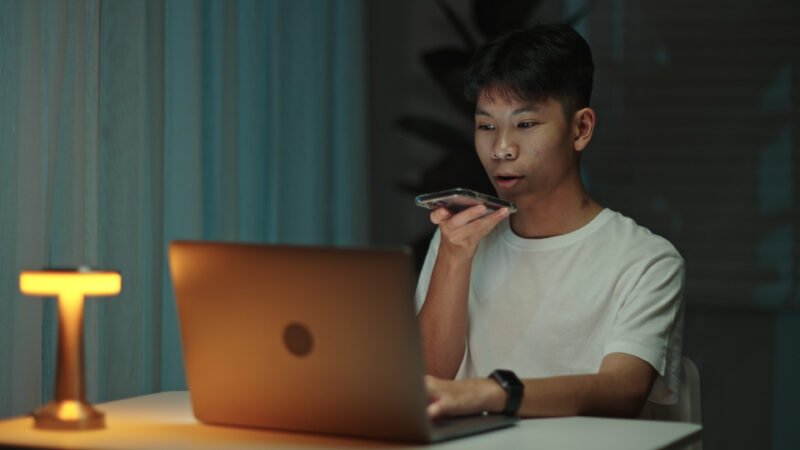 teenage boy student talking with a voice assistant on smartphone while working at home during night.