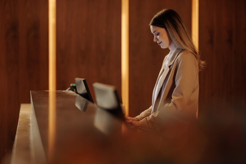 Young woman working at a hotel reception desk, engaging with guests while skillfully using a computer in a modern, cozy lobby setting