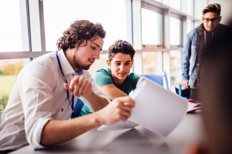 Teacher showing a student some results on his paperwork. The student looks happy as they sit side by side at a table. Another student looks on.
