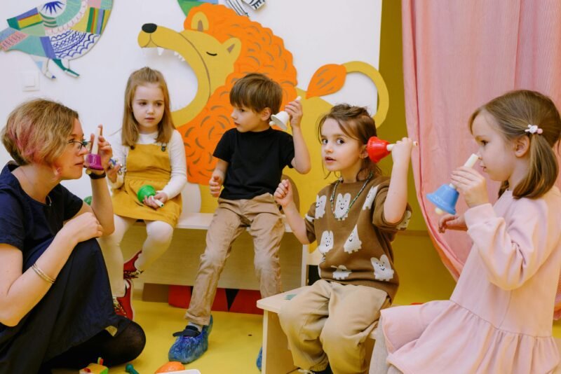 A teacher is kneeling on the floor surrounded by young students holding bells.