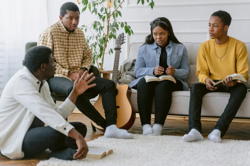 A man sits cross legged on the floor while three people sit on a couch or in a chair with a guitar beside them. 