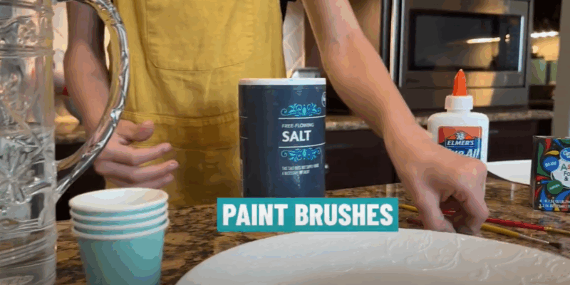 A child is seen from the neck down standing in front of a table that has cups, a plate, a pitcher, a container of salt, Elmer's glue, or food coloring. 