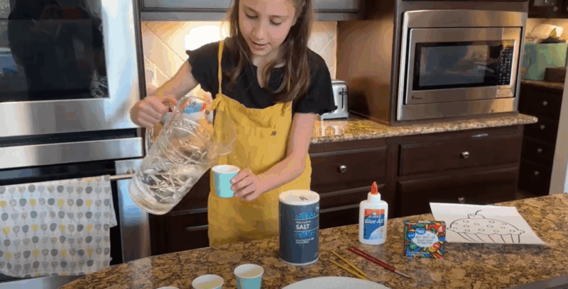 A little girl is seen pouring water into a small Dixie paper cup in this first step in how to make a salt painting.