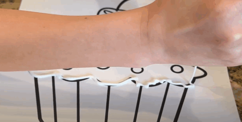A hand is seen squeezing white glue along the lines of a cupcake coloring page in this second step of salt painting.