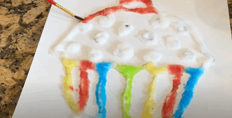 A cupcake coloring page has glue and salt spread on it. A paintbrush is shown spreading color onto it. 