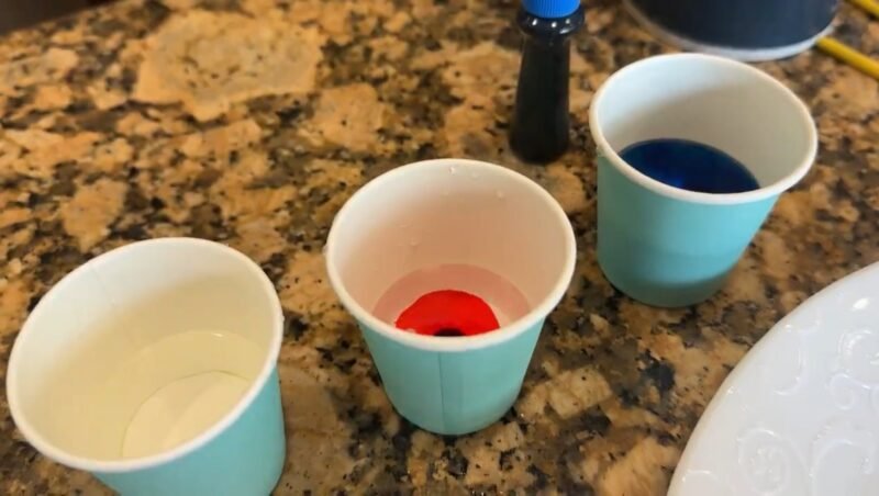 Several small paper cups are shown on a counter top filled with water. A drop of food coloring has been added into the cups in this second step of salt painting. 