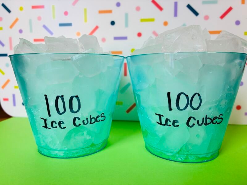 two blue cups with 100 mini ice cubes sitting in front of a rainbow background to conduct a melting ice cube experiment in the class to celebrate the 100th day of school